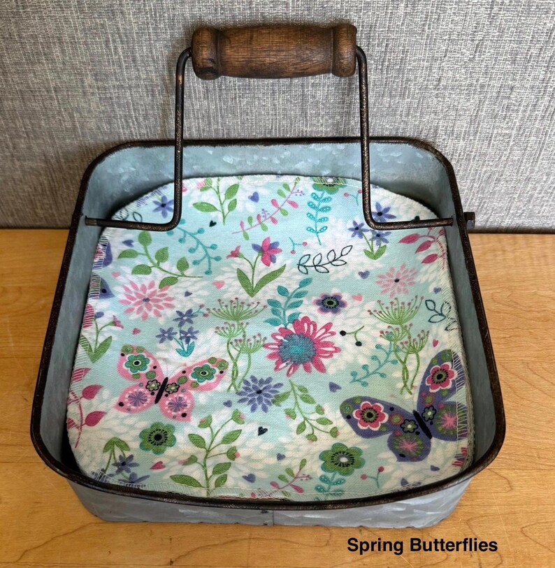May include: A square galvanized metal napkin holder with a dark brown wooden handle. Inside sits a stack of napkins featuring a light blue background with a colorful pattern of pink and purple butterflies, green leaves, and various floral designs. Text reads Spring Butterflies.