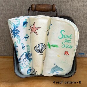 Pode incluir: Tr&ecirc;s panos de tecido dobrados com estampas marinhas, incluindo uma tartaruga, uma estrela-do-mar, uma concha e um golfinho. O pano da direita exibe o texto "Save our Seas" em verde. Os panos est&atilde;o em uma cesta de metal com al&ccedil;a de madeira.