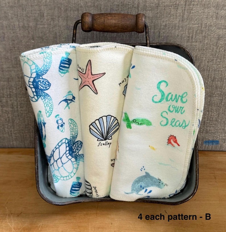 Pode incluir: Tr&ecirc;s panos de tecido dobrados com estampas marinhas, incluindo uma tartaruga, uma estrela-do-mar, uma concha e um golfinho. O pano da direita exibe o texto "Save our Seas" em verde. Os panos est&atilde;o em uma cesta de metal com al&ccedil;a de madeira.