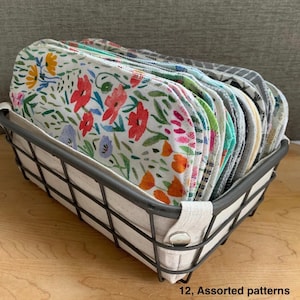 May include: A metal wire basket with a linen lining holds a stack of twelve assorted fabric pads. The pads feature various floral and patterned designs, including red, orange, and blue flowers on a white background. The text "12, Assorted patterns" is visible.