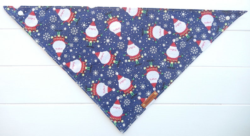May include: A blue bandana with a repeating pattern of Santa Claus figures and snowflakes. The bandana has a brown leather tag with the word "Charming" printed on it.
