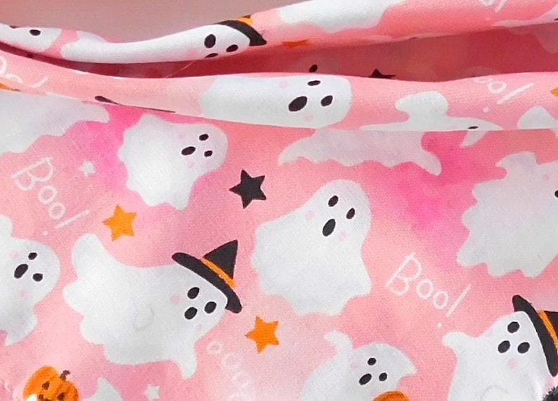 Puede incluir: Tela rosa con tem&aacute;tica de Halloween con fantasmas blancos, estrellas negras y estrellas y calabazas naranjas. Algunos fantasmas llevan sombreros de bruja. La palabra "Boo!" est&aacute; escrita en blanco en la tela.
