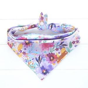 May include: A white bandana with a purple, pink, and yellow floral print. The bandana has a white button closure and a brown leather tag that says "Chumleys".