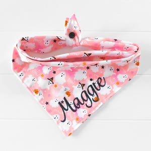 Puede incluir: Bandana rosa de Halloween para perros con un estampado de fantasmas y calabazas blancas. La bandana est&aacute; personalizada con el nombre "Maggie" en escritura negra. Cierre de bot&oacute;n.