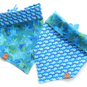 May include: Two blue dog bandanas with sea-themed patterns. One features turtles and starfish on a watercolor background, the other seahorses. Both have a contrasting seahorse pattern on the folded edge and a small leather tag.