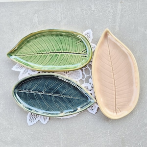 Op de afbeelding: Drie blad-vormige keramische schalen in groen, blauw en perzik. Elke schaal heeft een gedetailleerd bladnervenpatroon. De schalen zijn gerangschikt op een wit kanten kleedje, met een op de natuur geïnspireerd ontwerp.