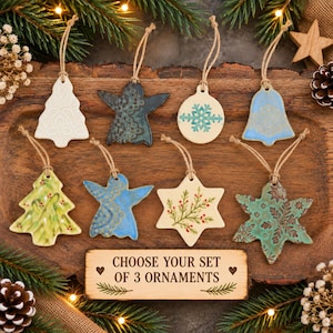 May include: A set of eight ceramic Christmas ornaments in various shapes including trees, stars, angels, a bell, and a snowflake. Each ornament features unique patterns, textures, and colours, suspended by twine loops. A wooden sign reads CHOOSE YOUR SET OF 3 ORNAMENTS.