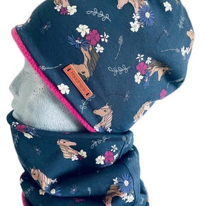 Op de afbeelding: Een marineblauwe muts en nekwarmer set met een bloemen- en paardenpatroon. De set heeft een roze rand en een klein leren label met de tekst "Handmade". Het patroon bevat witte, paarse en blauwe bloemen.