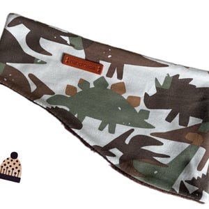 Op de afbeelding: Brede hoofdband met een dinosauruspatroon in bruin, groen en wit. Een klein bruin leren label met de tekst "Handmade". De stof is zacht en het ontwerp herhaalt zich over het hele oppervlak.