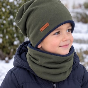 Könnte beinhalten: Eine olivgrüne Beanie-Mütze und ein passender Schlauchschal. Die Mütze hat ein kleines braunes rechteckiges Etikett mit dem in Schwarz aufgedruckten Wort Handmade. Beide Teile sind mit einem dunkelblauen, strukturierten Stoff gefüttert.