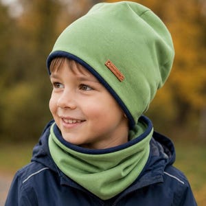 Puede incluir: Un conjunto de gorro y braga de cuello verdes con ribete azul marino. El gorro tiene una pequeña etiqueta de cuero marrón que dice "Handmade". El conjunto está hecho de un material suave y lo lleva un niño.