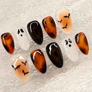 以下が含まれることがあります： ハロウィーンをテーマにしたつけ爪のセット。オレンジと茶色のマーブル模様、黒いクモ、白い幽霊、グラデーションの背景にコウモリのデザインが含まれています。爪は楕円形で、すぐに使用できます。