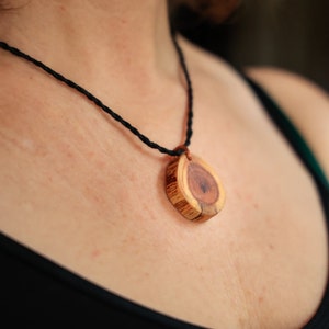 May include: A close-up of a wooden pendant necklace with a natural wood grain pattern. The pendant is round and has a dark brown centre. The necklace is made of black cord.