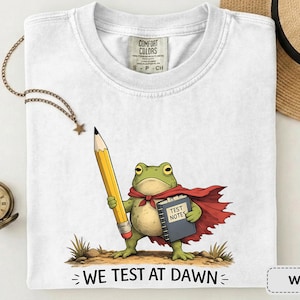 May include: White t-shirt with a cartoon frog wearing a red cape, holding a large yellow pencil and a book labelled "Test Notes." The text "WE TEST AT DAWN" is below the frog. A straw hat and a pocket watch are in the background.