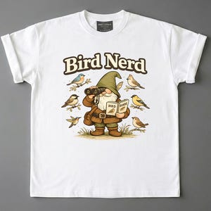 May include: A white t-shirt featuring a graphic of a gnome wearing a green hat and brown clothing, holding binoculars and a book labelled Bird Guide. The gnome is surrounded by six small birds of various colours, with the text Bird Nerd above the gnome.