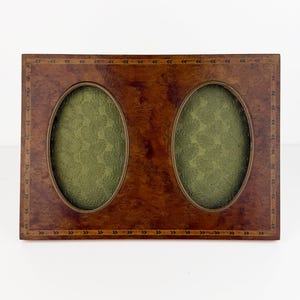 Puede incluir: Un marco de fotos doble rectangular con acabado en madera de raíz marrón y un borde incrustado decorativo. El marco presenta dos aberturas ovaladas, cada una con un inserto de tela verde oliva texturizada con un patrón de hojas.