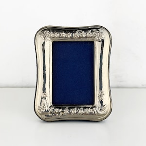 May include: A rectangular silver-toned picture frame with rounded corners and a polished metallic finish. The frame features an embossed floral pattern along the top and bottom edges, surrounding a deep navy blue rectangular insert.