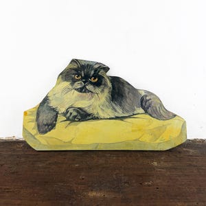 Könnte beinhalten: Ein flaches, ausgeschnittenes Deko-Objekt aus Holz mit einer gemalten Illustration einer langhaarigen Katze mit dunkelgrauem und cremefarbenem Fell, bernsteinfarbenen Augen und flachem Gesicht, die auf einem hellgelben Kissen ruht.