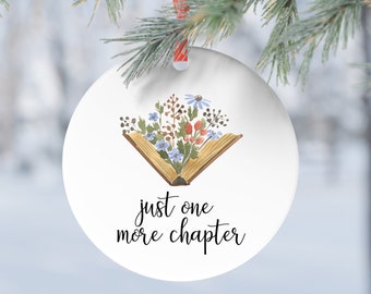May include: A white, round Christmas ornament with a watercolor illustration of an open book filled with flowers. The text "just one more chapter" is written below the book in a script font. The ornament hangs from a red ribbon.