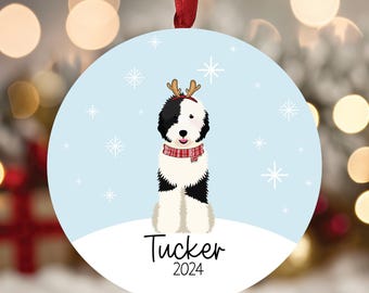 May include: Round Christmas ornament featuring a black and white dog wearing a red and white scarf and reindeer antlers. The dog is sitting on a snowy surface against a light blue background with snowflakes. The name "Tucker" and the year "2024" are printed below.