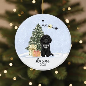 May include: A round ceramic Christmas ornament featuring a black doodle dog wearing a black and white buffalo plaid bandana. The scene includes a decorated Christmas tree, wrapped gifts, and Santa's sleigh in the sky. The text reads Bruno and 2026.