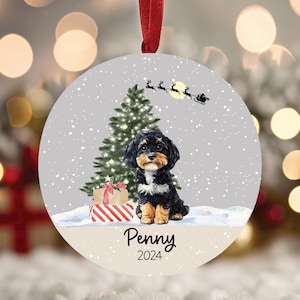 May include: A round Christmas ornament featuring a black and brown dog sitting in front of a decorated Christmas tree with a red and white striped gift box. The ornament has a gray background with falling snow and the text "Penny 2024".