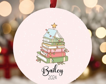 May include: A round, pale pink Christmas ornament featuring a stack of books arranged in a tree shape, topped with a yellow star and wrapped in string lights. The name Bailey and the year 2024 are printed in black script and sans-serif text below.