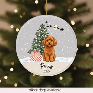 May include: A round ceramic Christmas ornament featuring a brown puppy sitting in snow in front of a green Christmas tree with gifts. Above the puppy, a silhouette of Santa and reindeer flies across a yellow moon. The text reads Penny and 2026.