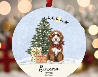 May include: Round Christmas ornament featuring a brown and white dog wearing a red and black plaid bandana. The ornament has a Christmas tree, presents, and a Santa sleigh scene. The name "Bruno" and the year "2025" are printed on the bottom.