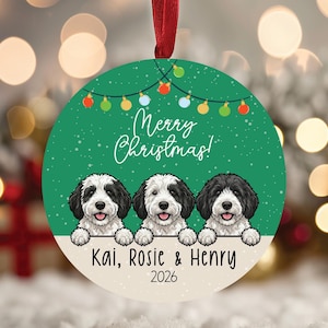 May include: A round green Christmas ornament featuring a string of colorful lights, white snowflakes, and three illustrated black and white puppies. The text reads Merry Christmas, Kai, Rosie & Henry, and 2026. It hangs from a red ribbon.