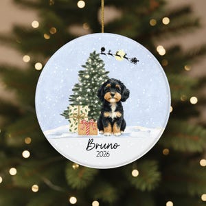 May include: A round ceramic Christmas ornament featuring a black and tan puppy sitting in front of a decorated Christmas tree and two gift boxes. Above, a silhouette of Santa and reindeer flies across a moon. The text reads Bruno and 2026.