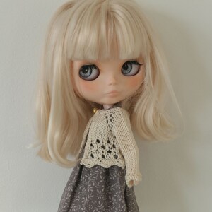 May include: A doll with long, light blonde hair and large blue eyes. The doll is dressed in a cream-coloured, open-weave sweater and a grey floral print dress. A collectible doll.