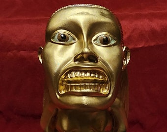 May include: A gold-colored statue of a snarling, human-like figure with a large mouth and teeth. The figure is kneeling on a black, circular base with intricate designs.