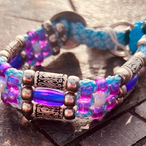 May include: A pet collar featuring a braided light blue cord interwoven with vibrant purple and iridescent beads. The design includes metallic silver spacers and patterned cylindrical beads, finished with a circular metal ring attachment.