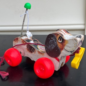 Op de afbeelding: Een vintage houten trekdier in de vorm van een hond met rode wielen, een gele plastic bot en een groene knop bovenop. De hond heeft een bruine neus en zwarte ogen. Het speelgoed is gelabeld met "Fisher Price Little People".