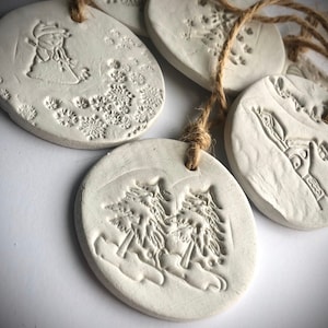 Puede incluir: Un conjunto de adornos redondos de arcilla blanca con diseños invernales en relieve que incluyen un muñeco de nieve, pinos y una casa, cada uno suspendido por un lazo de cuerda de yute natural con texturas detalladas en relieve.