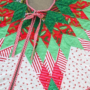 May include: A green, red, and white patchwork Christmas tree skirt with a red and white gingham ribbon tied in a bow. The skirt is made of various fabrics with different patterns, including stars, stripes, and polka dots.