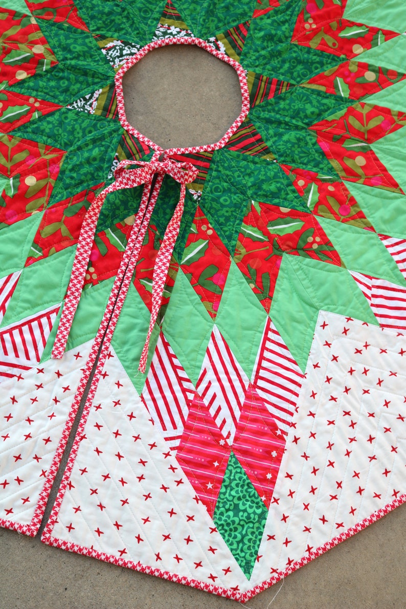 May include: A green, red, and white patchwork Christmas tree skirt with a red and white gingham ribbon tied in a bow. The skirt is made of various fabrics with different patterns, including stars, stripes, and polka dots.