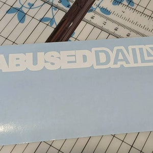 May include: A light blue sticker with the words "ABUSED DAILY" in white, bold, sans-serif font. The sticker is rectangular and appears to be on a cutting mat with a grid pattern.