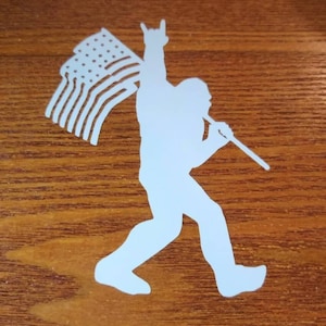May include: White silhouette of Bigfoot holding an American flag and making a rock and roll hand gesture. The image is set against a wood grain background.