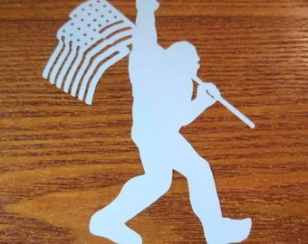 May include: White silhouette of Bigfoot holding an American flag and making a rock and roll hand gesture. The image is set against a wood grain background.