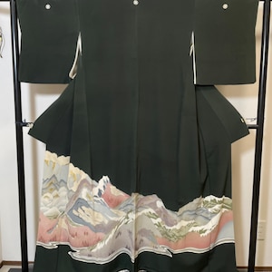 May include: A dark green kimono with a mountain landscape design at the hem. The kimono is displayed on a black rack. The mountain scene features shades of pink, gray, and white, creating a serene and artistic look.