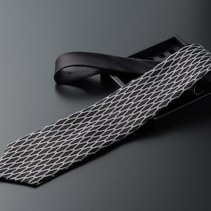 Puede incluir: Una corbata de patrón negro y blanco con un diseño geométrico. La corbata está doblada y colocada en una bandeja negra.