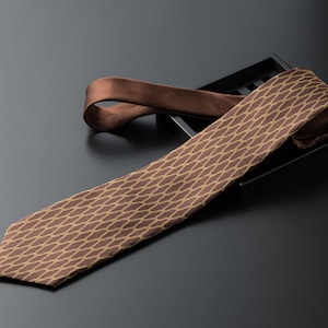 Puede incluir: Una corbata marrón con un patrón repetitivo de eslabones dorados. La corbata se muestra sobre una superficie negra, con el nudo en una bandeja negra. Un accesorio clásico para vestimenta formal o de negocios.