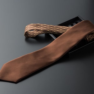 Puede incluir: Dos corbatas de seda marrones, una con un patrón geométrico, sobre una superficie negra.