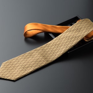 Puede incluir: Una corbata beige con un patrón de cadena repetido se muestra sobre una superficie negra. Una corbata naranja se coloca detrás de la corbata beige. Es probable que las corbatas estén hechas de seda o un material similar.