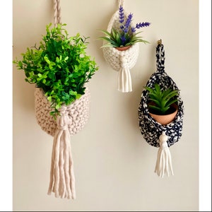 May include: Three hanging plant pots with macrame tassels. The pots are made of crocheted cotton yarn in white and black. The plants are a succulent, lavender, and a green leafy plant.