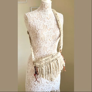 Puede incluir: Un bolso bandolera de crochet blanco con detalles de flecos y una correa de cuero marrón.
