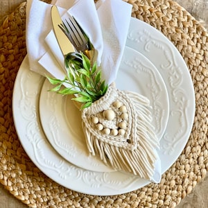 以下が含まれることがあります： 白いナプキンに、銀のフォークとナイフが、木製のビーズが付いたベージュの編み込みナプキンリングで包まれています。ナプキンリングはフリンジが付いたハート型です。ナプキンは白いプレートの上に置かれ、その下に白いプレートが置かれています。プレートは織りのプレースマットの上に置かれています。
