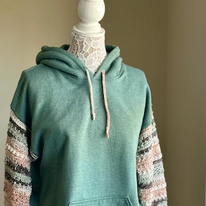 以下が含まれることがあります： 緑色のフード付きスウェットシャツ。袖は茶色、ピンク、白の糸で編まれたクロシェ編みです。